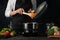 The professional chef in black apron pouring frying carrot from pan wok into pot with soup on dark blue background. Backstage of