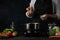 The professional chef in black apron salting water before cooking soup on dark blue background. Backstage of preparing meal.