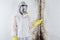 A professional disinfector in overalls and a mask points a finger at the black mold on the wall. Removal of the fungus in the apar