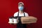 Professional food delivery man in a protective mask and gloves holds boxes of pizza and coffee and gives the client, fast delivery