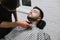 Professional hairdresser does a beard hairstyle to a client with a trimmer in his hands, lies with his eyes closed on a chair in a