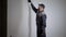 Professional painter is wearing working uniform and cap is painting wall by roller with white color, side view