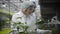 Professional senior Caucasian woman in protective workwear adding fertilizer to seedlings in glasshouse. Young man