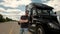 Professional truck driver approaches his truck and crosses his arms behind him parked long haul semi-truck with cargo