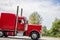 Profile of bright classic red big rig semi truck tractor with vertical chrome pipes running on the green highway