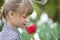 Profile of cute pretty smiling child girl with gray eyes and long hair smelling bright red tulip flower on blurred sunny summer