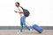 Profile portrait of young female traveler walking with cellphone and suitcase against white wall