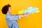 Profile side photo of young dark skin woman shoot water gun empty space isolated on yellow color background