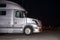 Profile of silver big rig semi truck with turned on headlight standing on dark night parking lot