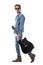 Profile view of cool confident man in jeans with sunglasses walking and carrying guitar looking behind.