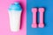 Protein cocktail bottle and pink dumbbells on pink and blue background, a top view