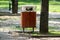 Public trash can made of narrow wooden boards with shiny metal top cover next to old tall tree surrounded with stone tiles in