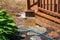 Puddle of brown water with soap suds on a brick patio near a brown wooden deck. There is water coming off of the deck.