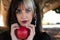 Punky girl with blonde and brunette hair holds a red apple in her hands and stares at it. The girl is a witch from the fairy tale