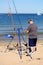 Purbeck, Dorset, UK - Jun 02 2018: Sea angler checking his tackle while on a sandy beach in the sunshine, with sailing boats on th