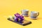 Purple crocuses lie on the phone against the background of two gray coffee cups, a yellow background, a place for text - the
