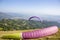 The purple green parachute wing lies on a hillside against the background of a paraglider taking off into a mountain valley