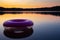 Purple inflatable swimming ring on top of still lake water surface at sunset. Rubber rescue wheel and colourful evening sky and