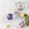 Purple tea in cup, teapot, lemons and blooming lilac on light background. Spring tea drinking. Greeting card, invitation design.