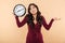 Puzzled brunette woman with curly long hair holding clock showing time after 8 gesturing like she is late or do not care over pea