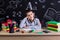 Puzzled excellent schoolboy sitting on the desk with books, school supplies, with one arm leaned under the cheeck