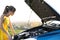 Puzzled female driver standing near her car with popped up hood inspecting broken engine