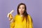 Puzzled pretty young woman in fur sweater blowing lips holding in hand fresh ripe banana fruit isolated on violet pastel