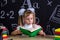 Puzzled schoolgirl sitting at the desk reading the book, surrounded with school supplies. Chalkboard as a background
