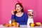 Puzzled woman keeps to healthy diet, bites fresh carrot, makes smoothie with food processor, wears red headband, blue sweater,