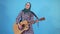 Puzzled young Muslim woman clumsily plays guitar on blue background