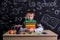 Puzzled youngster sitting at the desk with a pile of books under the chin, surrounded with school supplies. Chalkboard