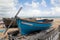 A quaint small blue wooden rowing boat berthed on wooden beams on a pebble beach next to another wood boat.