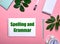 Question SPELLING AD GRAMMAR is written in green on a white notepad on a pink background surrounded by notepads, pens, white alarm