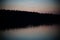 A quiet blue-reddish night sky at dusk with the silhouette of the forest is reflected in the dark forest lake when it rains