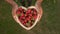 Rack focus of young woman holding a heart shaped wooden bowl of fresh strawberries outside