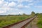 A railroad running diagonally across a green field in a countryside against a blue sky with clouds. Summer day