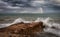 A rainbow in the dark clouds over the Mediterranean Sea with rocks in the foreground.