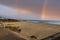 A rainbow over the beach with vast silky brown sand with people walking down the bike trail and people standing in the sand