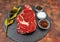 Raw beef ribeye on a round slate plate with glass containers containing peppercorns  coarse salt and cayenne chillies on an old