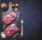 Raw beef steak with rosemary,butter and meat fork on dark rustic cutting board, top view with space for tex