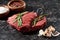 Raw beef steak with rosemary near garlic cloves and small bowls with salt and peppercorns on black marble surface.