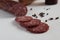 A raw cured stick of sausage and sausage slicing pieces lies on a white table to black pepper peas and pepper shakers