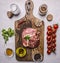 Raw pork in marinade, on a cutting board with tomatoes on a branch, oil, black pepper, herbs on wooden rustic background top view