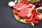 Raw ribeye beef steak ready for grilling with seasonings and vegetables on wooden cutting board over dark concrete background
