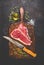 Raw T-bone Steak for grill or BBQ with fresh herbs and oil and kitchen knife on dark aged cutting board