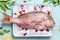 Raw whole fish in tray with ice cubes on light turquoise background with heabs and spices, top view. Seafood concept.