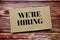 We`re Hiring written on paper card top view of wooden background