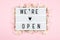 We`re open lettering on a lightbox on a pink background surrounded by flowers