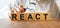 REACT inscription on the texture of wooden cubes. A business man holds a magnifying glass in his hand and examines the inscription