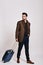 Ready to travel. Stylish dark-haired man standing with suitcase and looking away isolated over grey background